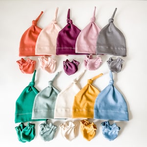 May include: A set of ten baby hats and ten pairs of baby mittens in various colours, including coral, pink, purple, grey, green, blue, cream, and yellow. The hats are tied at the top with a knot. The mittens are ruffled at the wrist.