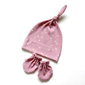 May include: A pink baby hat with a white cross pattern and matching mittens. The hat has a knot on top.