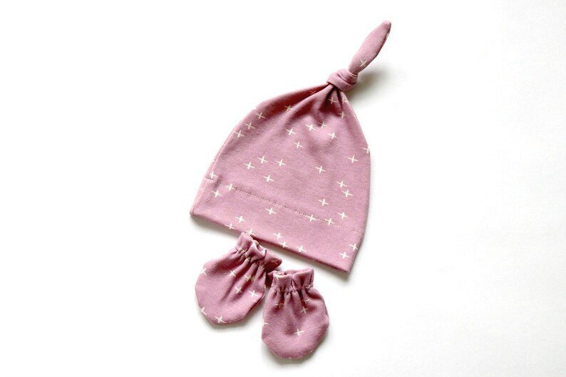 May include: A pink baby hat with a white cross pattern and matching mittens. The hat has a knot on top.