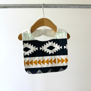 May include: A white, blue, and yellow patterned bib with a geometric design. The bib is hanging on a wooden hanger.