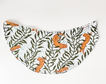 May include: A white fabric bib with a green and orange pattern of sea otters and seaweed. The bib has a white trim and two white snaps.