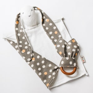 May include: A white hooded towel with brown and white polka dot bunny ears and a matching brown and white polka dot teether with a wooden ring. The towel has the text "B. Toys" on the bottom right corner.