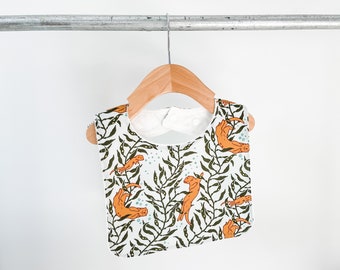 May include: A white baby bib with a green and orange print of sea otters swimming in seaweed. The bib is hanging on a wooden hanger.