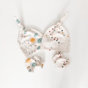May include: A set of baby accessories in a cream colour with a sun, moon, and cloud pattern. The set includes a knotted hat and two pairs of mittens.