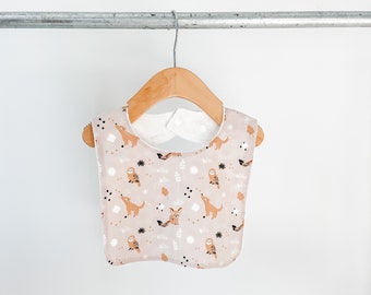 May include: A beige baby bib with a white trim and a woodland animal print featuring owls and foxes. The bib is hanging on a wooden hanger.