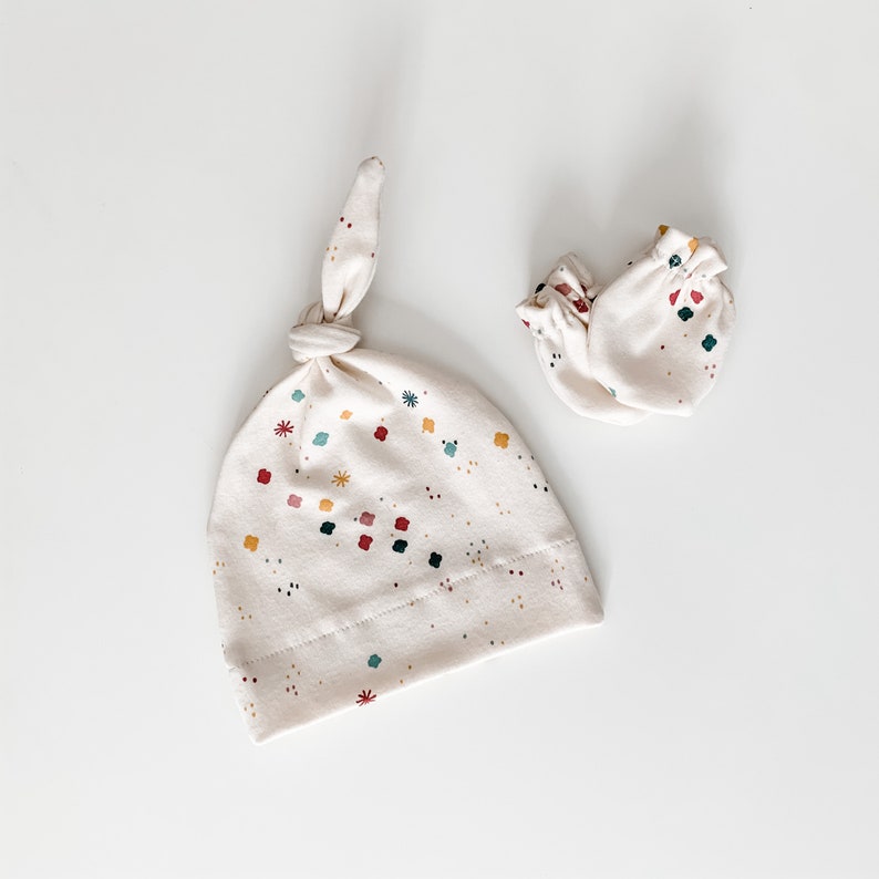May include: A set of baby accessories in a cream colour with a confetti print. The set includes a knotted hat and a pair of mittens.