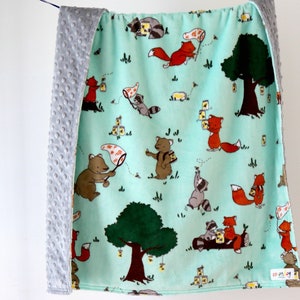 Puede incluir: Una suave manta de bebé de color verde menta con estampado de animales del bosque. La manta presenta zorros, mapaches y osos recogiendo miel en un entorno de bosque. La manta tiene un respaldo de minky gris.