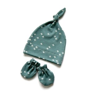 May include: A set of baby accessories in a teal blue color with a white cross pattern. The set includes a knotted hat and a pair of mittens.