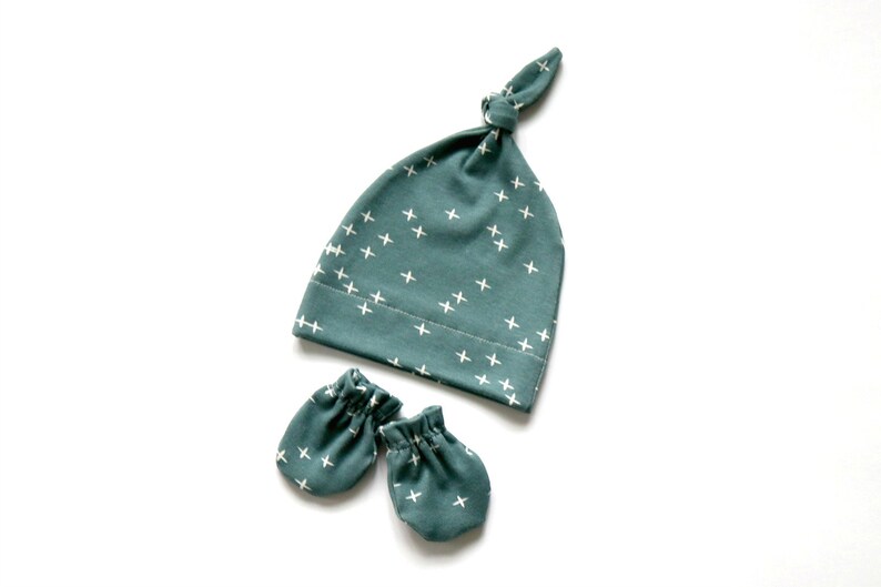 May include: A set of baby accessories in a teal blue color with a white cross pattern. The set includes a knotted hat and a pair of mittens.