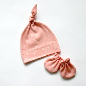 May include: A pink baby hat with a white plus sign pattern and matching mittens. The hat is tied at the top with a knot.