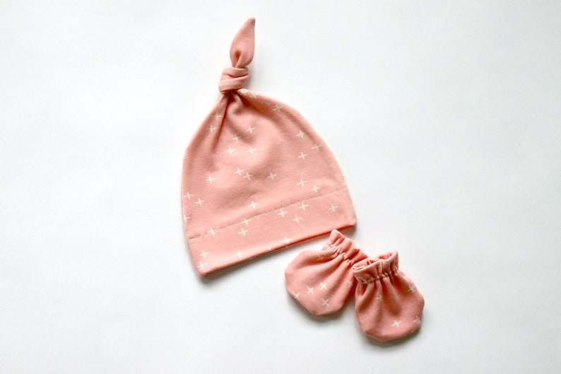 May include: A pink baby hat with a white plus sign pattern and matching mittens. The hat is tied at the top with a knot.