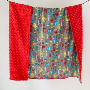 Può includere: Una copertina per neonati rossa e grigia con un motivo di robot colorati su un lato e un tessuto minky a pois rosso sull'altro.