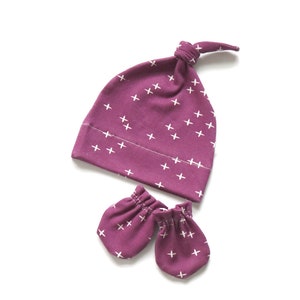 May include: A set of baby accessories in a deep purple color with a white cross pattern. The set includes a knotted beanie and a pair of mittens.