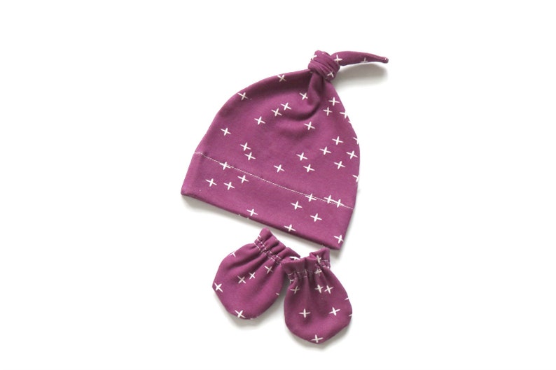 May include: A set of baby accessories in a deep purple color with a white cross pattern. The set includes a knotted beanie and a pair of mittens.
