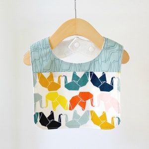 May include: A blue and white patterned baby bib with a white background and colorful origami cranes. The bib has a white fleece lining and two white snaps.