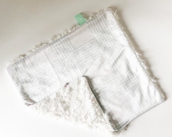 Puede incluir: Una manta de bebé de cuadros blancos y grises con un lado blanco suave y esponjoso. La manta tiene una etiqueta verde.