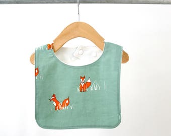 May include: A green baby bib with a white lining and a fox print. The bib is hanging on a wooden hanger.