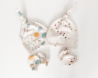 May include: A set of baby accessories in a cream color with a sun, moon, and cloud pattern. The set includes a knotted hat and two pairs of mittens.