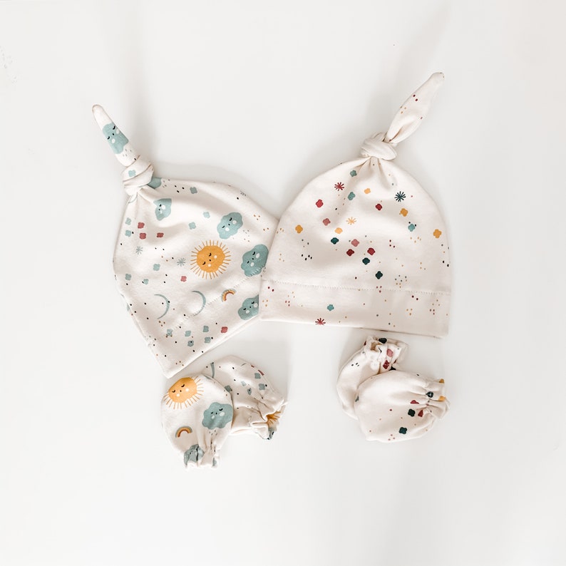 May include: A set of baby accessories in a cream colour with a sun, moon, and cloud pattern. The set includes a knotted hat and two pairs of mittens.