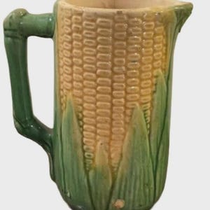 May include: A ceramic pitcher with a corn cob design. The pitcher features a yellow and tan corn cob pattern on the body, with green leaves at the base. It has a green handle and spout.