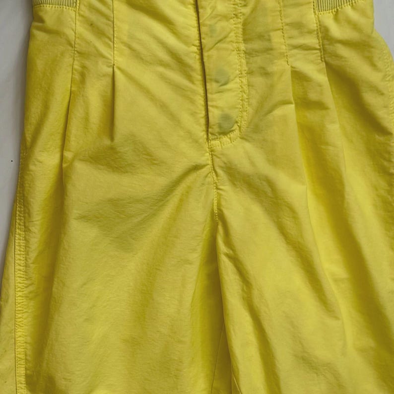 Puede incluir: Primer plano de unos pantalones cortos de color amarillo brillante. Los pantalones cortos tienen cintura alta, pliegues y cierre de bot&oacute;n. La tela parece ser un material ligero, con costuras visibles y una superficie texturizada. Los pantalones cortos son de color s&oacute;lido y vibrante.