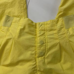 Puede incluir: Primer plano de unos pantalones de color amarillo brillante. Los pantalones tienen un frente plisado con detalles de costura verticales. La cintura presenta secciones el&aacute;sticas a cada lado. Hay manchas visibles en la parte delantera.