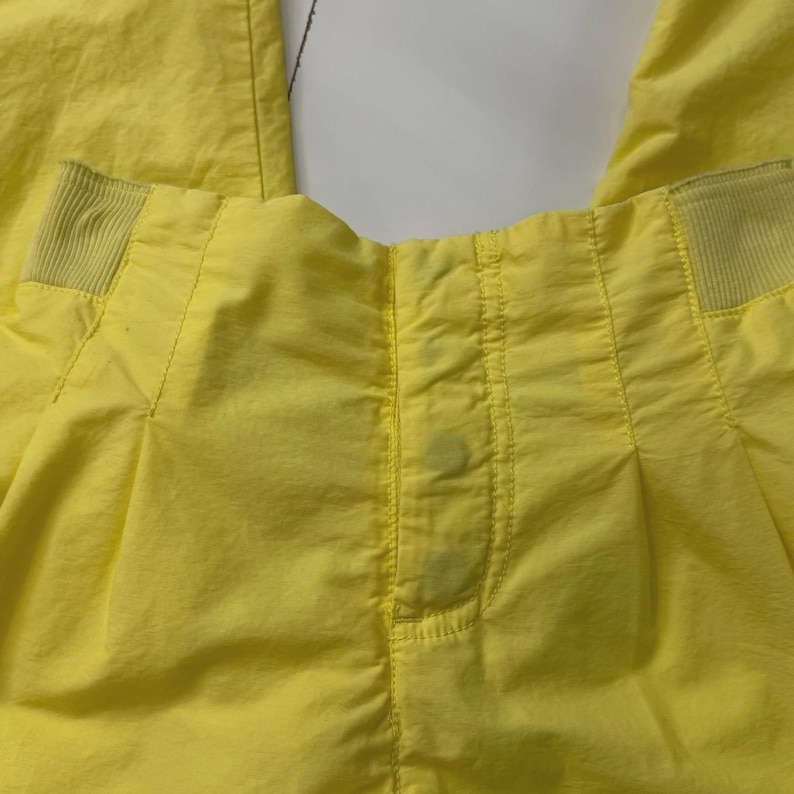 Puede incluir: Primer plano de unos pantalones de color amarillo brillante. Los pantalones tienen un frente plisado con detalles de costura verticales. La cintura presenta secciones el&aacute;sticas a cada lado. Hay manchas visibles en la parte delantera.