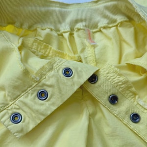 Puede incluir: Primer plano de una prenda amarilla con cierres de botones a presi&oacute;n met&aacute;licos. La tela es de un amarillo claro liso, con costuras visibles y una cintura acanalada. Los botones a presi&oacute;n son de color plateado con un centro oscuro.