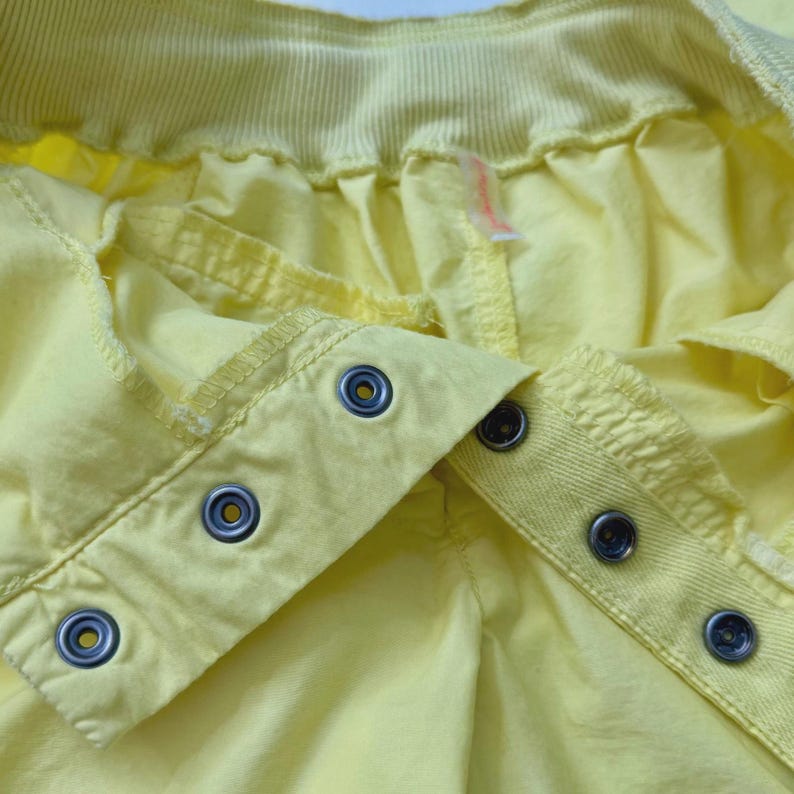 Puede incluir: Primer plano de una prenda amarilla con cierres de botones a presi&oacute;n met&aacute;licos. La tela es de un amarillo claro liso, con costuras visibles y una cintura acanalada. Los botones a presi&oacute;n son de color plateado con un centro oscuro.