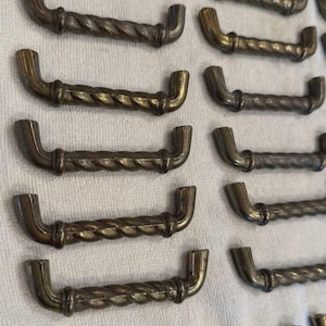 May include: A collection of antique bronze-colored cabinet handles. Each handle features a twisted rope design in the center and curved ends. The handles are arranged on a white surface, showcasing their vintage aesthetic.