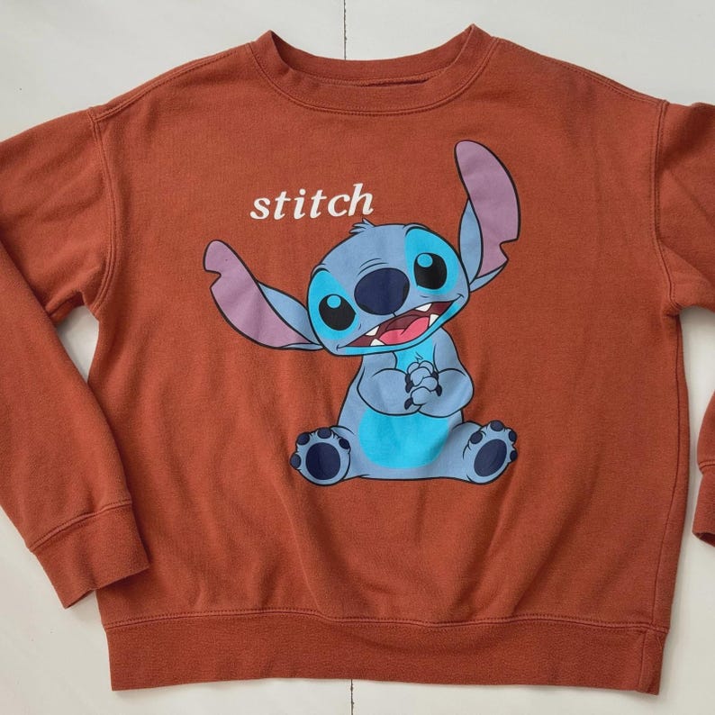 Puede incluir: Sudadera de color &oacute;xido con una imagen de dibujos animados de Stitch, una criatura extraterrestre azul. La palabra "stitch" est&aacute; impresa encima del personaje. La sudadera tiene cuello redondo y mangas largas.