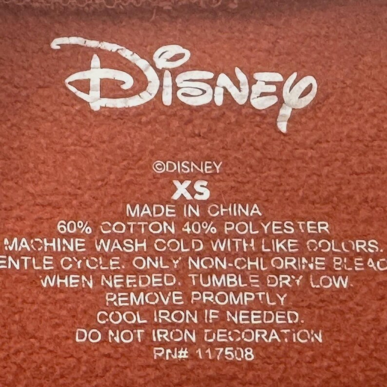 Puede incluir: Primer plano de una etiqueta de tela marr&oacute;n rojizo con el logotipo de Disney en blanco. La etiqueta incluye texto que indica que la prenda est&aacute; hecha en China, es talla XS y es 60% algod&oacute;n y 40% poli&eacute;ster. Tambi&eacute;n se imprimen instrucciones de lavado.