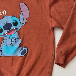 Puede incluir: Sudadera de color &oacute;xido con un gran gr&aacute;fico de dibujos animados de Stitch de Lilo & Stitch. El personaje es azul con ojos grandes, una gran sonrisa y orejas grandes. La sudadera tiene cuello redondo y mangas largas.