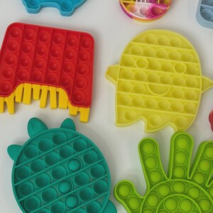 Puede incluir: Una colecci&oacute;n de coloridos juguetes fidget pop-it en varias formas. Incluye un rect&aacute;ngulo rojo con detalles amarillos, un fantasma amarillo, una tortuga verde y una mano verde. Estos juguetes de silicona est&aacute;n dise&ntilde;ados para ser presionados y reventados.