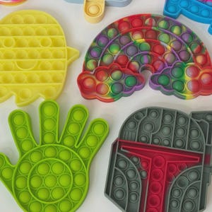 Puede incluir: Una colecci&oacute;n de juguetes sensoriales de silicona coloridos en varias formas. Incluye un fantasma amarillo, un arco&iacute;ris, una mano verde y un casco gris. Cada juguete presenta m&uacute;ltiples depresiones en forma de burbujas que se pueden empujar hacia adentro y hacia afuera.