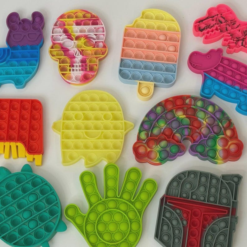 Puede incluir: Una colecci&oacute;n de coloridos juguetes fidget pop-it en varias formas, incluyendo una llama, una calavera, un helado, un dinosaurio, un hipop&oacute;tamo, un rect&aacute;ngulo, un fantasma, un arco&iacute;ris, una tortuga, una mano y un casco. Los juguetes tienen burbujas multicolores.