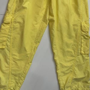 Puede incluir: Pantalones cargo de color amarillo brillante con m&uacute;ltiples bolsillos. Los pantalones est&aacute;n hechos de un material ligero y tienen un ajuste relajado. La imagen muestra un primer plano de los pantalones, destacando los detalles de los bolsillos y las costuras.