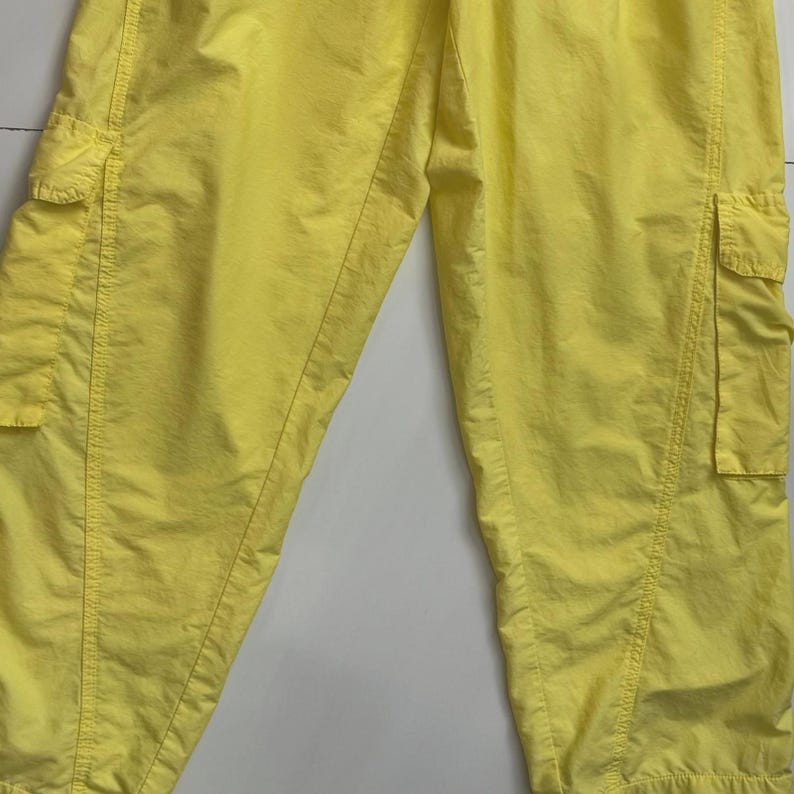 Puede incluir: Pantalones cargo de color amarillo brillante con m&uacute;ltiples bolsillos. Los pantalones est&aacute;n hechos de un material ligero y tienen un ajuste relajado. La imagen muestra un primer plano de los pantalones, destacando los detalles de los bolsillos y las costuras.