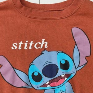 Puede incluir: Sudadera de color &oacute;xido con cuello redondo que presenta un personaje de dibujos animados con detalles en azul, morado y negro. La palabra "stitch" est&aacute; impresa en blanco. El personaje tiene orejas grandes y una amplia sonrisa.