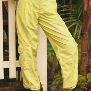 Puede incluir: Pantalones cargo de color amarillo brillante con m&uacute;ltiples bolsillos y un ajuste relajado. Los pantalones se combinan con zapatos blancos sin cordones. La imagen est&aacute; tomada al aire libre, con una valla blanca y vegetaci&oacute;n en el fondo.