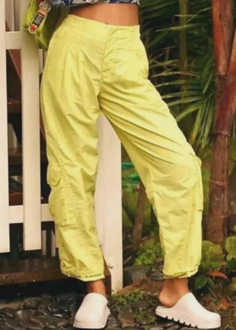 Puede incluir: Pantalones cargo de color amarillo brillante con m&uacute;ltiples bolsillos y un ajuste relajado. Los pantalones se combinan con zapatos blancos sin cordones. La imagen est&aacute; tomada al aire libre, con una valla blanca y vegetaci&oacute;n en el fondo.