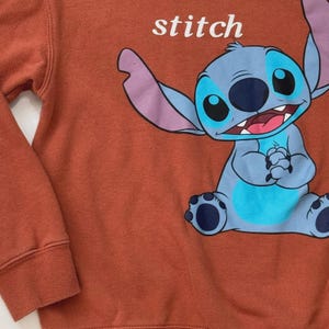 Puede incluir: Sudadera de color &oacute;xido con un gran gr&aacute;fico del personaje Stitch de la pel&iacute;cula de Disney Lilo & Stitch. La palabra "stitch" est&aacute; impresa en blanco encima del personaje. El personaje es azul con grandes ojos negros y orejas moradas.