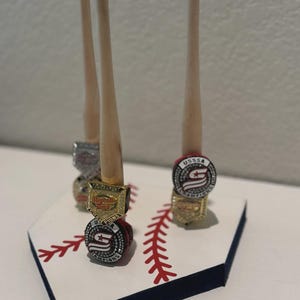 May include: A baseball-themed display stand featuring wooden dowels and championship rings. The base is white with red stitching and a blue border, resembling a baseball home plate. The rings are gold and silver.
