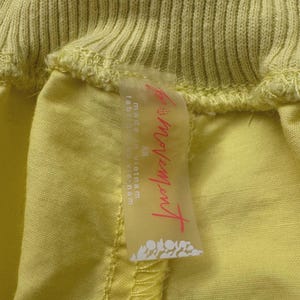 Puede incluir: Primer plano de una prenda amarilla clara con cintura acanalada. Una etiqueta transparente est&aacute; adjunta, con el texto "Movement" en escritura rosa, junto con "made in Vietnam" y "XS" en negro. La tela de la prenda es de color amarillo p&aacute;lido.