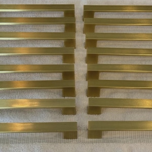 Könnte beinhalten: Eine Sammlung von messingfarbenen Schrankgriffen, angeordnet auf einer weißen Oberfläche. Jeder Griff hat ein rechteckiges Stangendesign mit quadratischer Basis. Die Griffe sind gleichmäßig angeordnet.