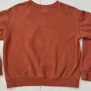 Puede incluir: Una sudadera de cuello redondo de color &oacute;xido. La parte superior de manga larga tiene cuello, pu&ntilde;os y dobladillo acanalados. La tela parece ser un material suave y de peso medio. La sudadera es de color s&oacute;lido sin gr&aacute;ficos ni patrones visibles.