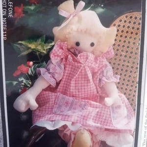 May include: A handmade doll with a light yellow hat tied with a pink bow. The doll wears a pink gingham dress with white lace trim and small pockets. The text "PICK ME UP - CARRY ME" is visible at the top of the image.