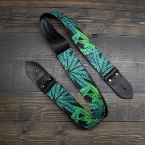 Puede incluir: Una correa de guitarra negra con un patrón de hojas de marihuana verdes. La correa tiene dos extremos de cuero ajustables.