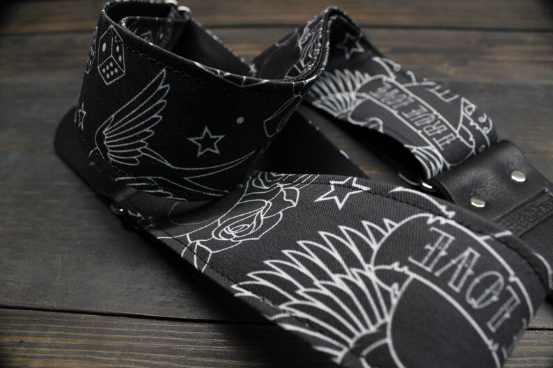 May include: Black guitar strap with white tattoo-style designs. The strap features images of a swallow, roses, stars, dice, and banners with the words "TRUE LOVE" and "LOVE". The strap has black leather ends and metal rivets.
