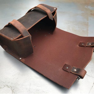 May include: A brown leather motorcycle saddlebag with two straps and buckles. The bag is open and the inside is lined with brown leather.